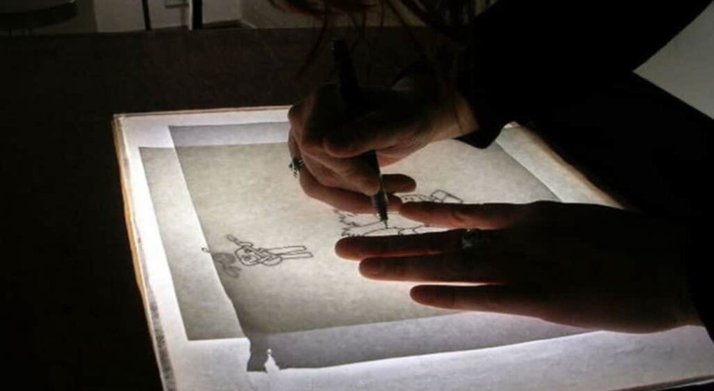 An artist's hands tracing over a sketch on a lightbox